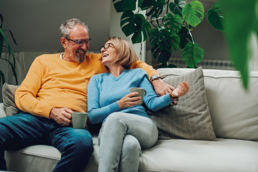 An image of an older couple looking happy, sitting on a sofa with cups of tea.