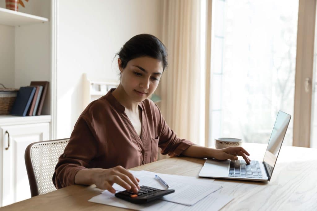 An image of a woman using a calculator while working on a laptop.