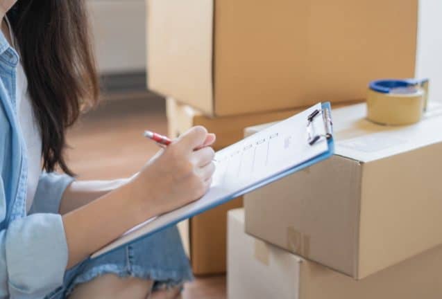 An image of a woman surrounded by boxes with a clipboard and parcel tape.